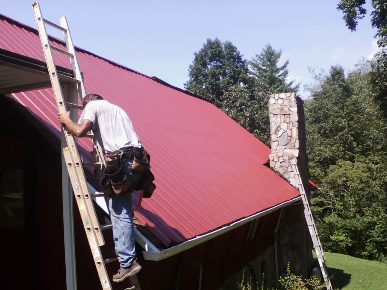 Roofer on a ladder beside a red metal roof and stone chimney — Soffit & Fascia Repair by Roofing Brothers in Wytheville